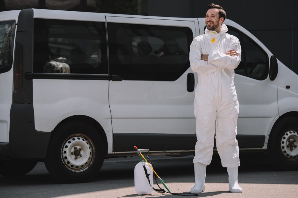 smiling pest control worker in uniform standing with crossed arms near car and sprayer on street